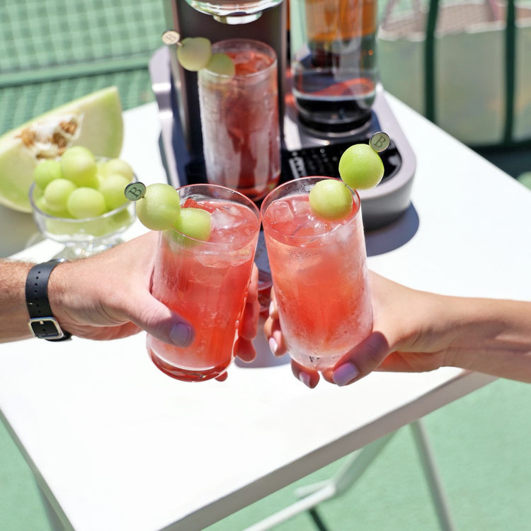 Two hands holding glasses of iced Golden Ace with honeydew garnish. A Bartesian cocktail maker and sliced melon are visible in the background.
