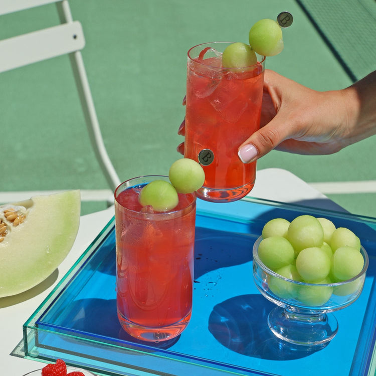 Two tall glasses of Golden Ace with honeydew garnish, set on a translucent blue tray with a bowl of melon balls, with a hand holding one glass against a green outdoor tennis court background.